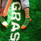 Lifestyle shot of a person wearing the 'Home Is Where The Weed Is' socks, sitting on a green shag carpet. [lf]