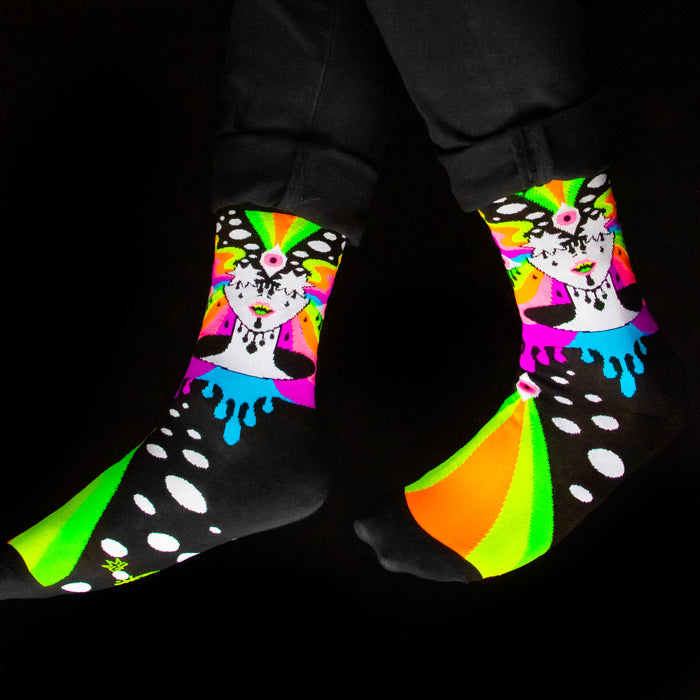 A pair of black socks with a colorful pattern of a woman's face with rainbow eyes and dripping paint. The socks are being worn by a person with dark pants.