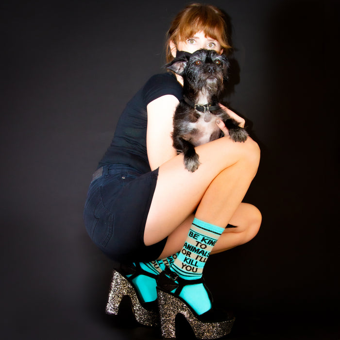 A young woman is crouching on the ground with a small dog in her arms. The woman is wearing a black shirt, blue sparkly shoes, and denim shorts. The dog is black and white and has a collar on. The woman has her hands around the dog and is looking at the camera.