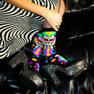 A person is wearing a pair of black and silver glitter platform shoes, black and white striped dress, and rainbow psychedelic socks with skulls on them. The person is sitting on a black leather couch.