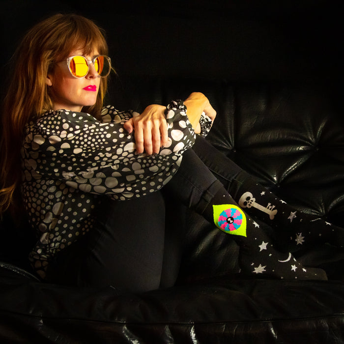 A woman with long red hair is sitting on a black leather couch. She is wearing a black-and-white polka dot blouse, black pants, and yellow sunglasses. She is also wearing a pair of socks with skulls and stars on them. The woman is looking down at her feet.