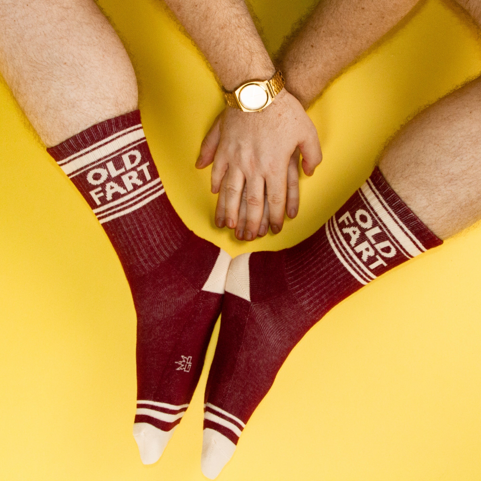 Flatlay shot showing two pairs of 'Old Fart' socks, with hands clasped in the center, against a yellow background. [lf]