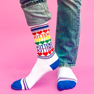 A person is modeling a pair of white socks with a colorful pattern of rainbow stripes and red heart-shaped outlines of the word 
