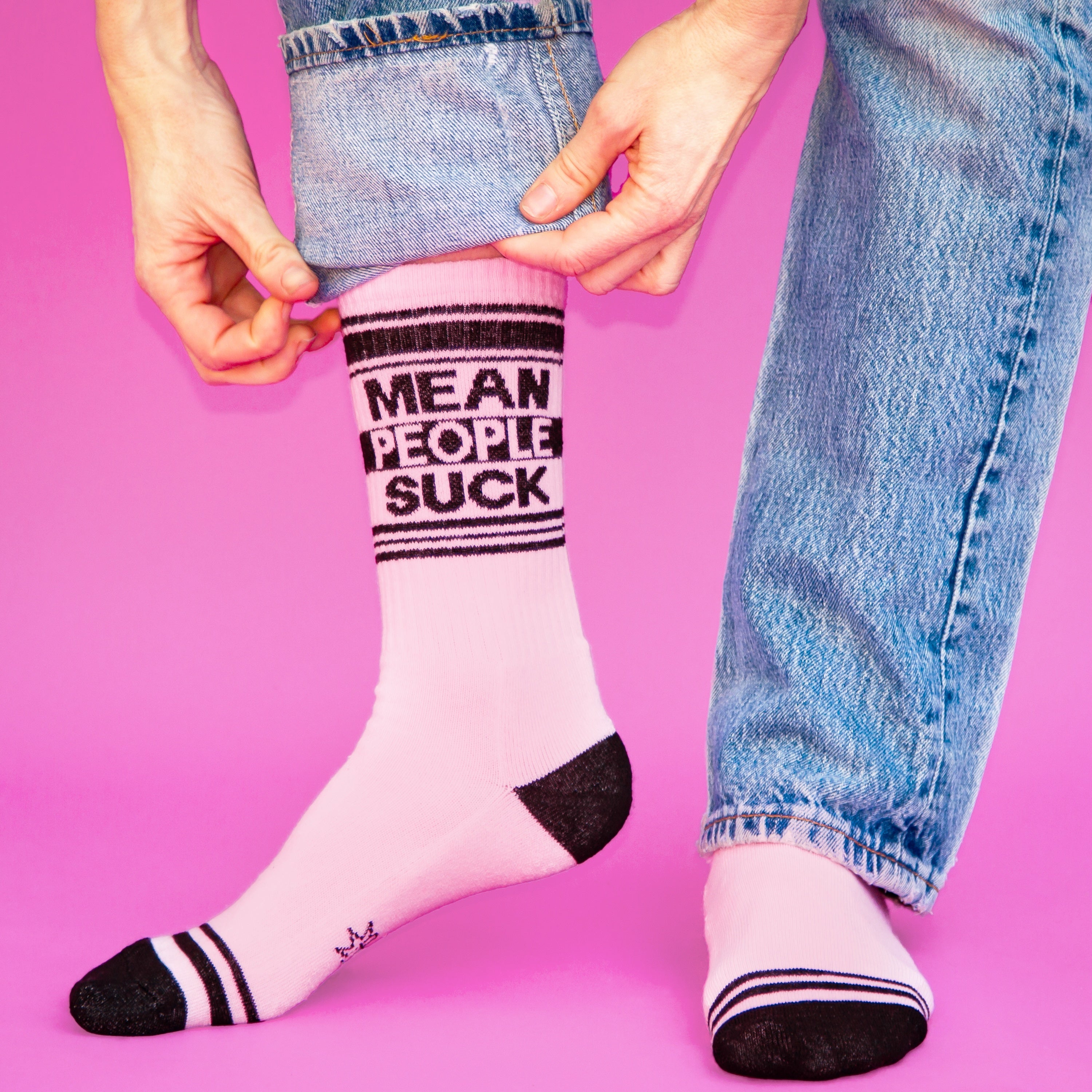 Lifestyle photo: Person wearing the 'Mean People Suck' socks with jeans, shown from the knees down on a pink background. [lf]