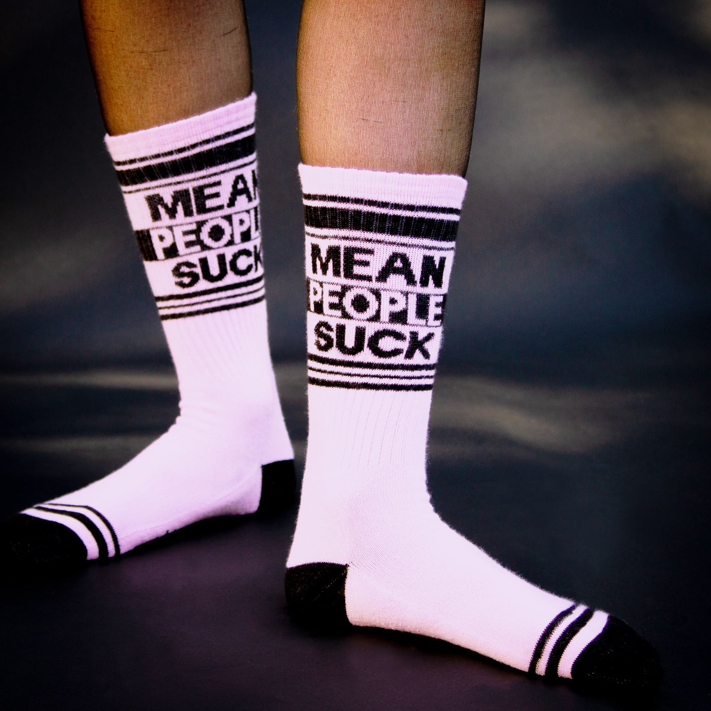 Close-up product shot of a person wearing a pair of pink 'Mean People Suck' socks. The socks are shown from the ankles down [lf]
