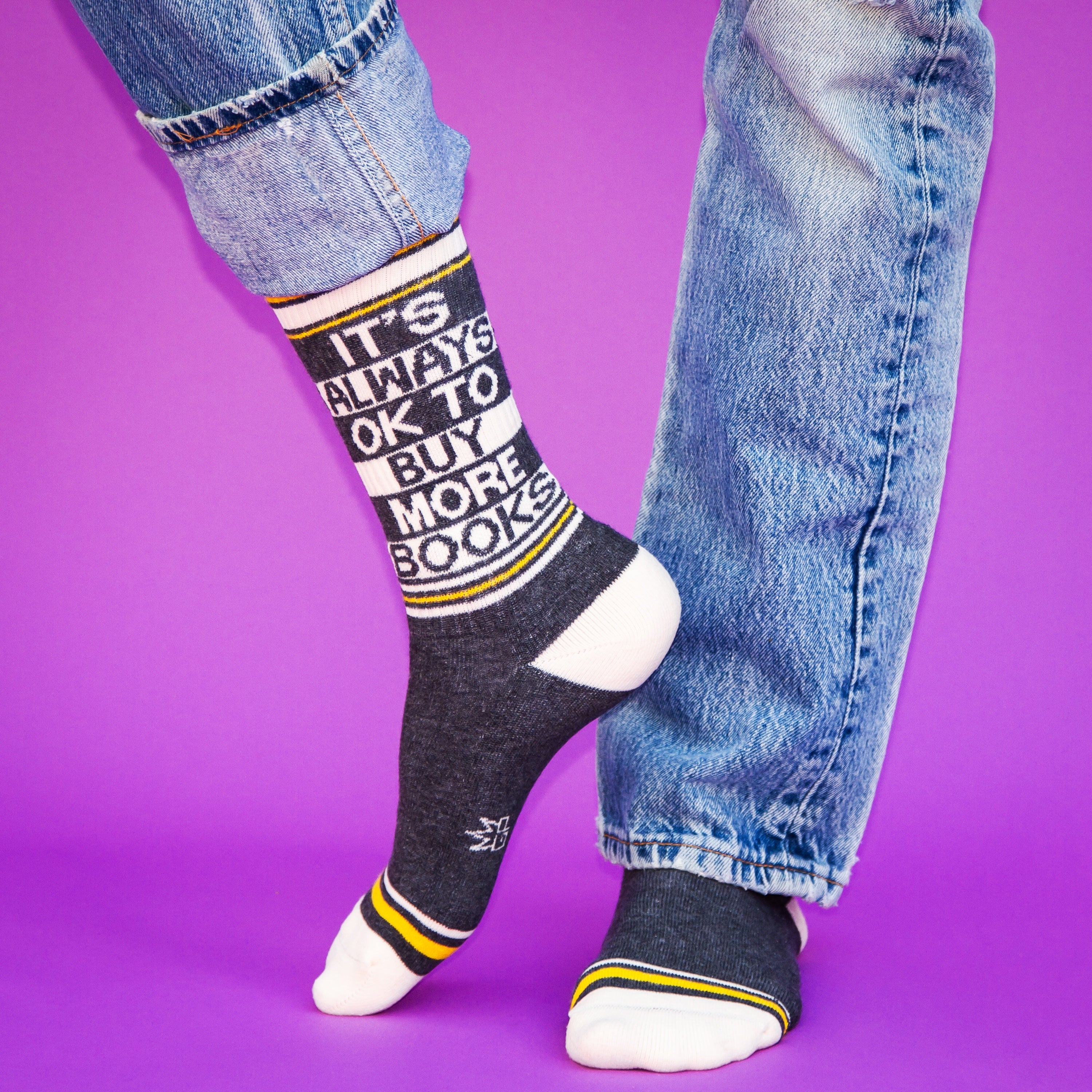 Close-up flatlay product shot of a pair of 'It's Always Ok To Buy More Books' socks against a purple background. [lf]