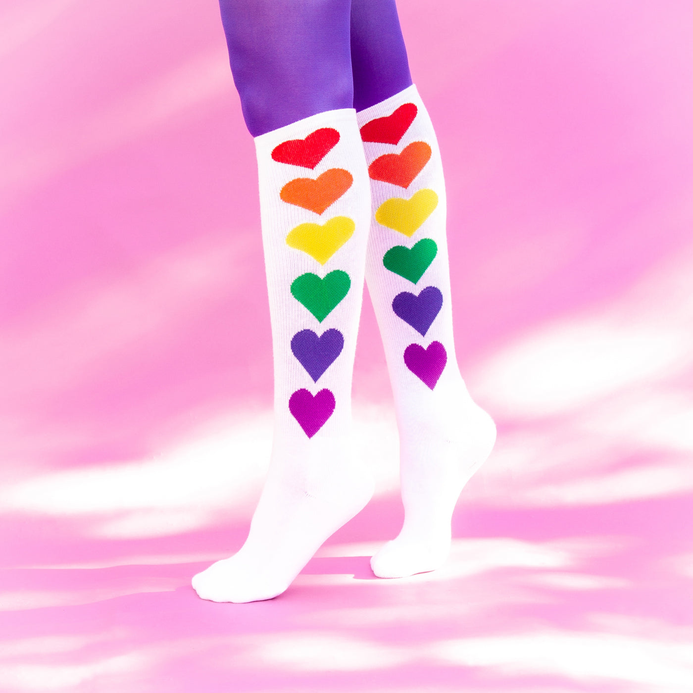 Lifestyle photo: Rainbow Hearts socks on a model's legs against a pink background. [lf]