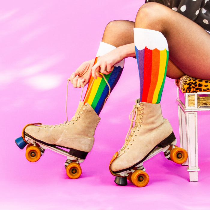 A pair of legs is shown from the knees down. The person is wearing purple tights and rainbow socks that have yellow toes and white cuffs. The socks have a pattern of thin and thick stripes in the colors red, orange, yellow, green, blue and purple.