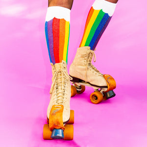 A pair of legs is shown from the knees down. The person is wearing purple tights and rainbow socks that have yellow toes and white cuffs. The socks have a pattern of thin and thick stripes in the colors red, orange, yellow, green, blue and purple.