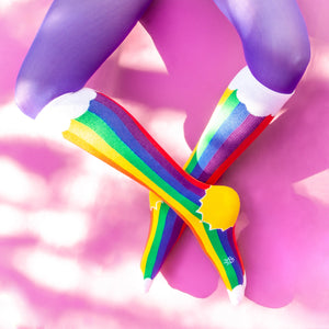 A pair of legs is shown from the knees down. The person is wearing purple tights and rainbow socks that have yellow toes and white cuffs. The socks have a pattern of thin and thick stripes in the colors red, orange, yellow, green, blue and purple.