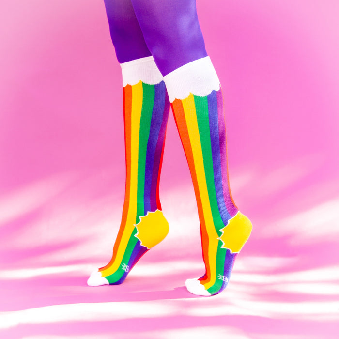 A pair of legs is shown from the knees down. The person is wearing purple tights and rainbow socks that have yellow toes and white cuffs. The socks have a pattern of thin and thick stripes in the colors red, orange, yellow, green, blue and purple.