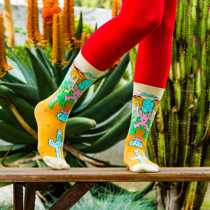 A person is sitting on a burnt orange fur style rug with their legs crossed. They are wearing mid-calf socks with a cactus pattern and white sneakers. The person is holding a pink rotary dial telephone receiver in their right hand.