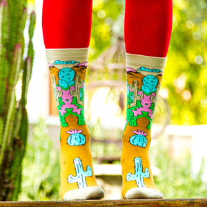 A person is sitting on a burnt orange fur style rug with their legs crossed. They are wearing mid-calf socks with a cactus pattern and white sneakers. The person is holding a pink rotary dial telephone receiver in their right hand.