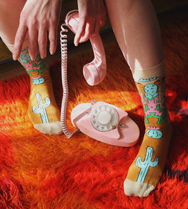 A person is sitting on a burnt orange fur style rug with their legs crossed. They are wearing mid-calf socks with a cactus pattern and white sneakers. The person is holding a pink rotary dial telephone receiver in their right hand.