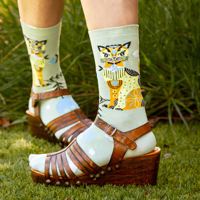 A pair of green socks with a colorful cat wearing a blue scarf design on the left sock and a pattern of blue and white clouds, yellow and blue birds, and black tree branches on both socks.