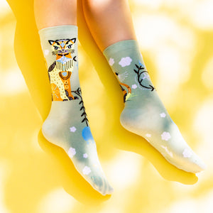 A pair of green socks with a colorful cat wearing a blue scarf design on the left sock and a pattern of blue and white clouds, yellow and blue birds, and black tree branches on both socks.