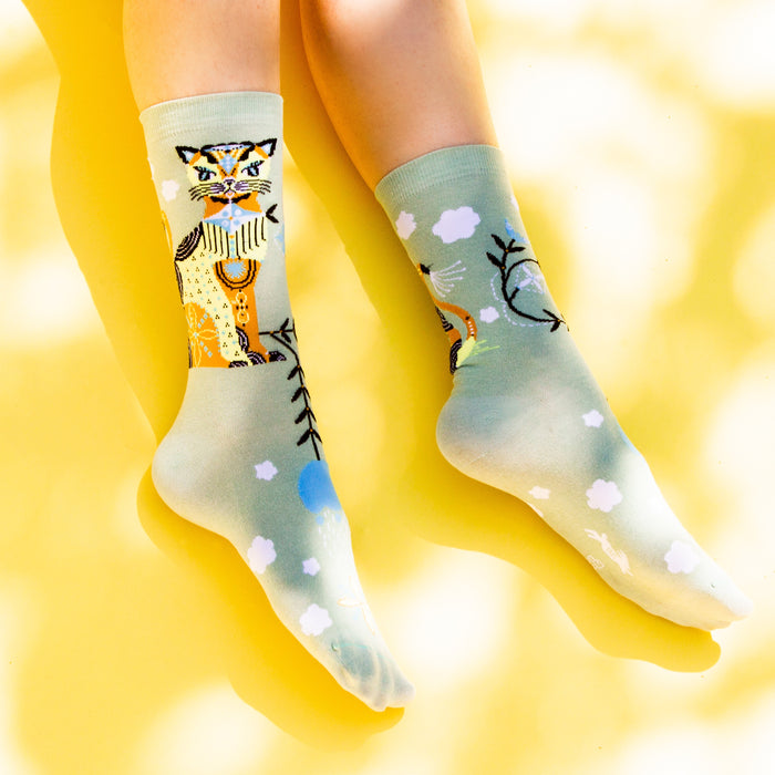 A pair of green socks with a colorful cat wearing a blue scarf design on the left sock and a pattern of blue and white clouds, yellow and blue birds, and black tree branches on both socks.