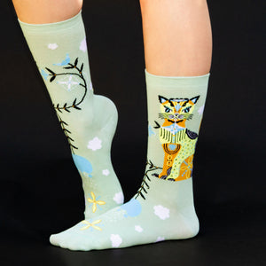 A pair of green socks with a colorful cat wearing a blue scarf design on the left sock and a pattern of blue and white clouds, yellow and blue birds, and black tree branches on both socks.