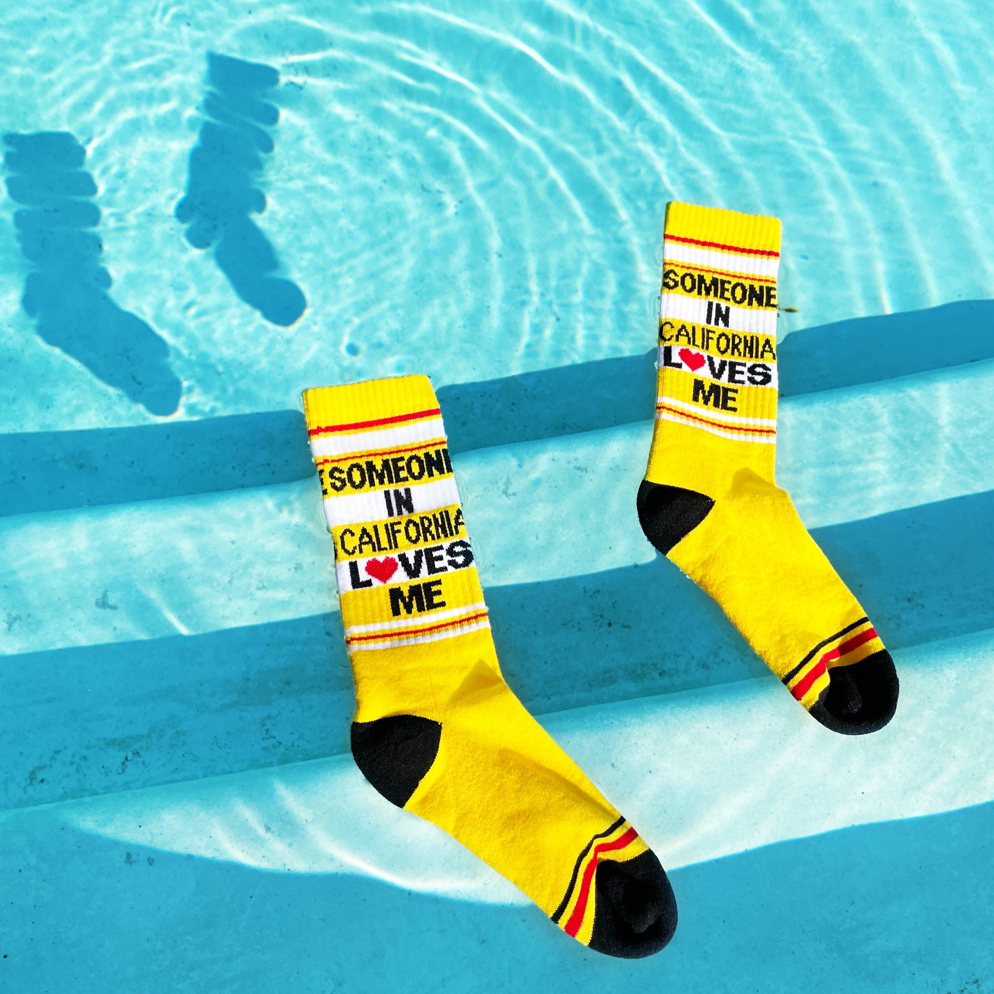 Lifestyle photo of yellow 'Someone in California Loves Me' socks resting on the edge of a pool. [lf]