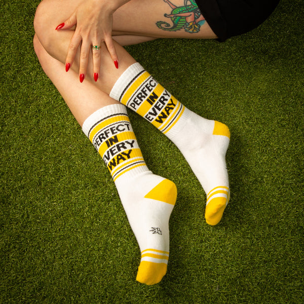 Lifestyle shot of a person's legs wearing the 'Perfect In Every Way' socks. The socks are white with yellow stripes and text. [lf]