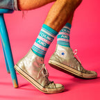 Lifestyle shot: Person wearing teal 'Young, Broke and Fabulous' socks with silver Converse shoes against a pink background. [lf]