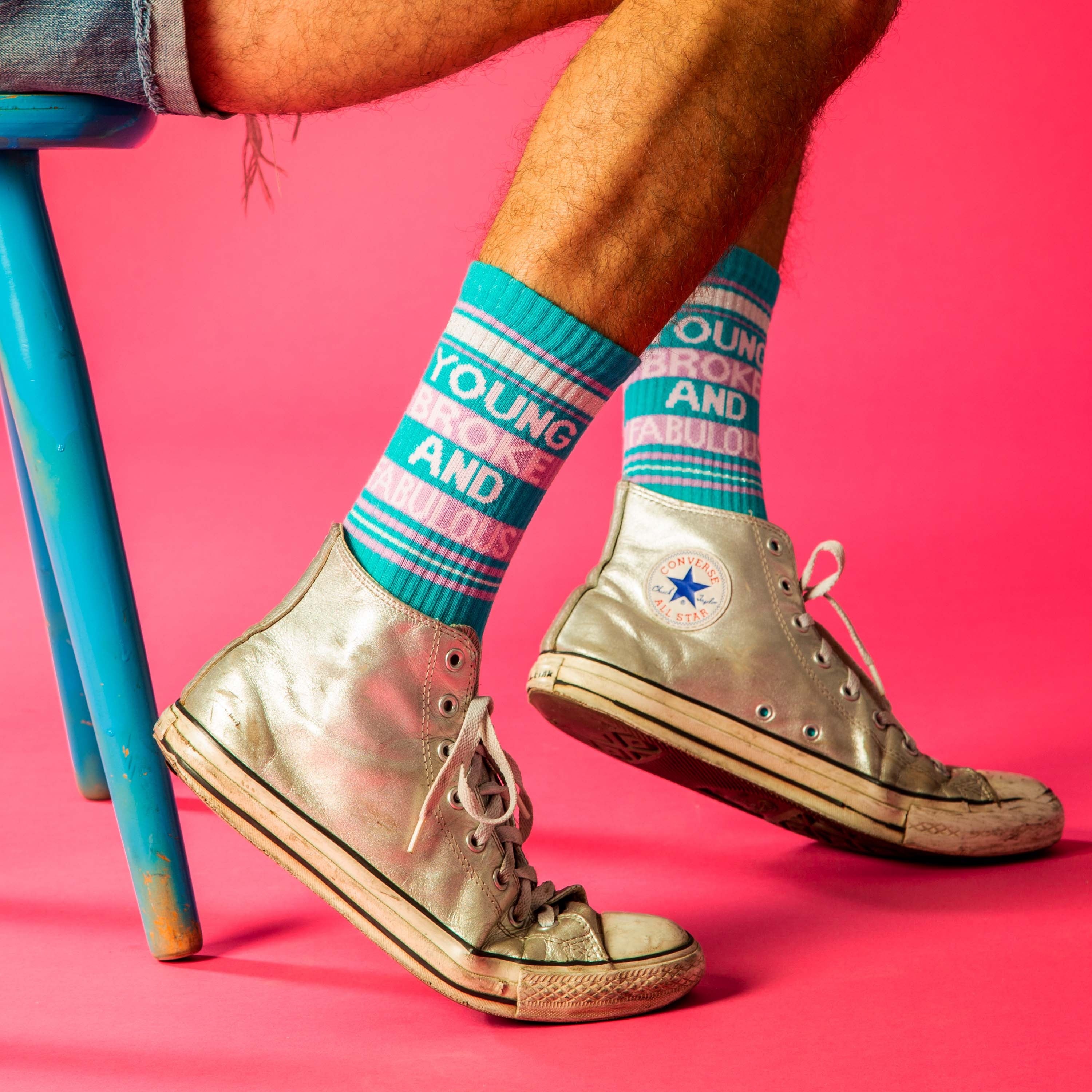 Lifestyle shot: Person wearing teal 'Young, Broke and Fabulous' socks with silver Converse shoes against a pink background. [lf]