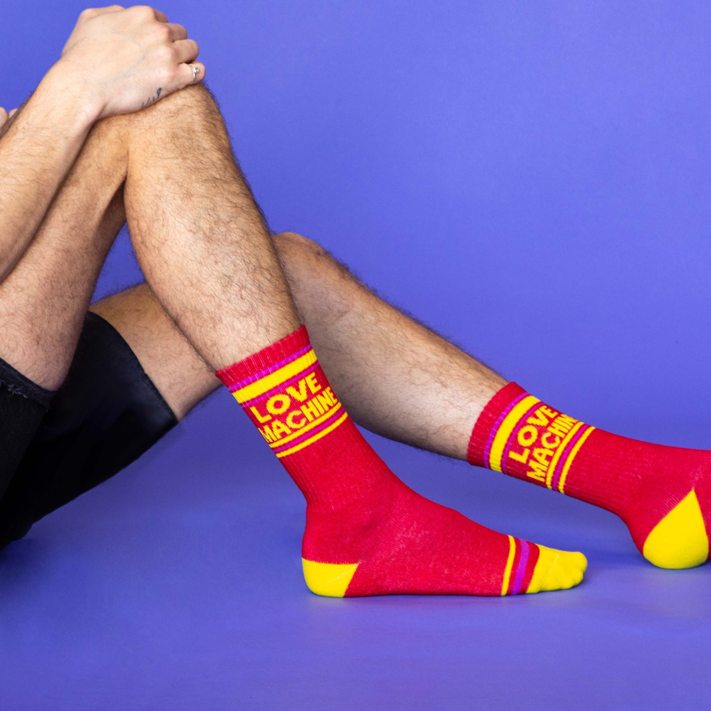 Lifestyle shot: Man wearing red 'Love Machine' crew socks, sitting on purple background. [lf]
