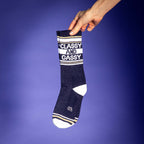 Close-up of a hand holding a single Classy and Gassy sock against a purple background. [lf]