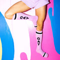 Lifestyle shot of a person's legs wearing the lavender 'GAY' socks against a colorful painted wall. [lf]