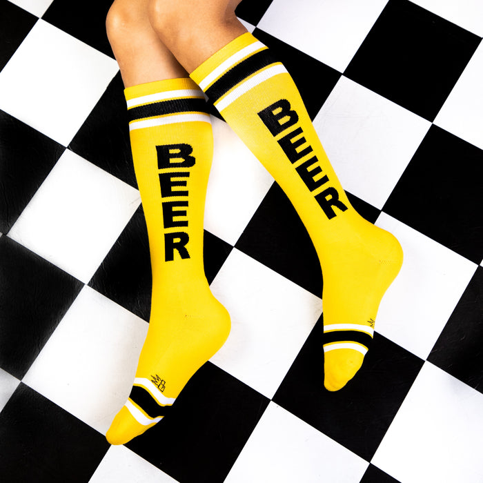 A pair of yellow socks with the word BEER in black letters. The socks are being worn by a person and their feet are on a black and white checkered floor.