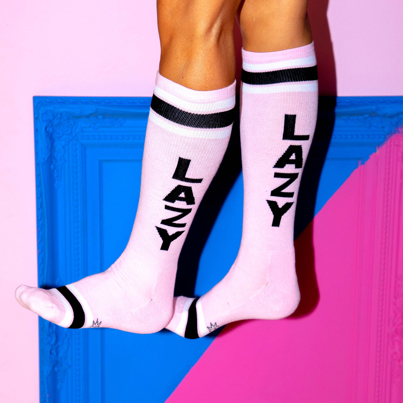 A fun lifestyle shot of 'Lazy' socks on a person's legs against a vibrant pink and blue backdrop. [lf]
