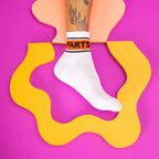 Lifestyle shot: A person's foot wearing a white 'Farts' sock on a vibrant pink and yellow background. [lf]