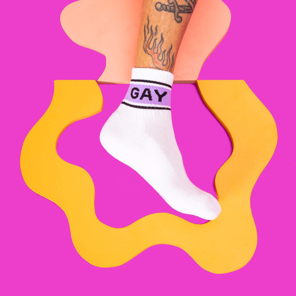 Close-up of a person's leg wearing one of the 'Gay' socks against a vibrant pink and orange background. [lf]