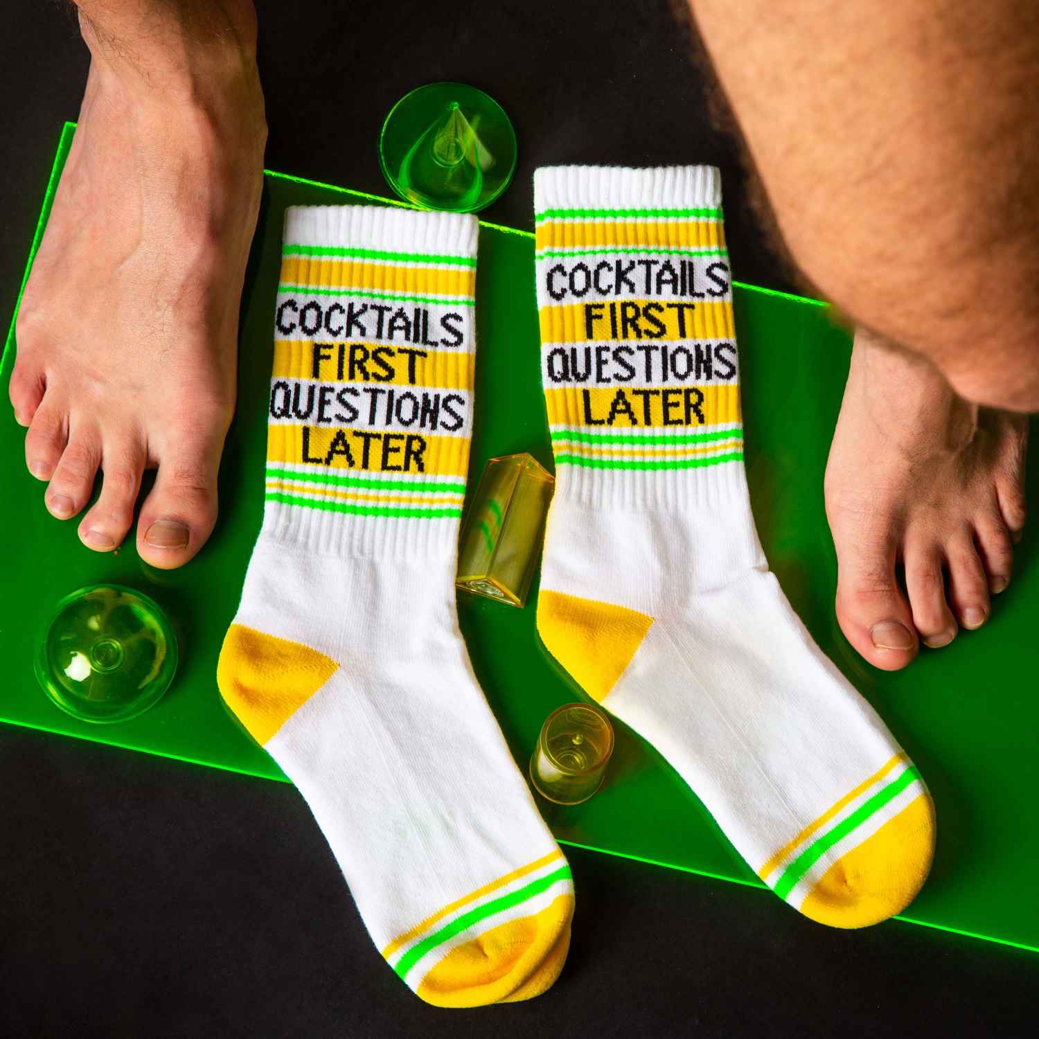 Flat lay of the 'Cocktails First Questions Later' socks on a bright green surface with small green objects. [lf]