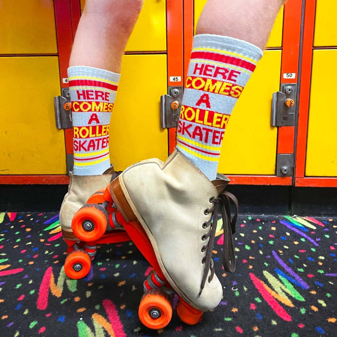 Close-up lifestyle shot: A person's feet in roller skates, showcasing the 'Here Comes A Roller Skater' socks against yellow lockers. [lf]