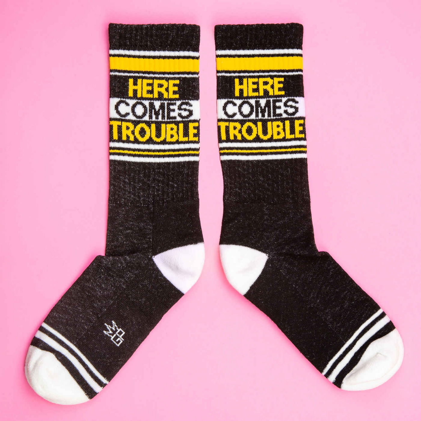 Another flat lay of two 'Here Comes Trouble' socks on a pink background; socks are arranged slightly differently.