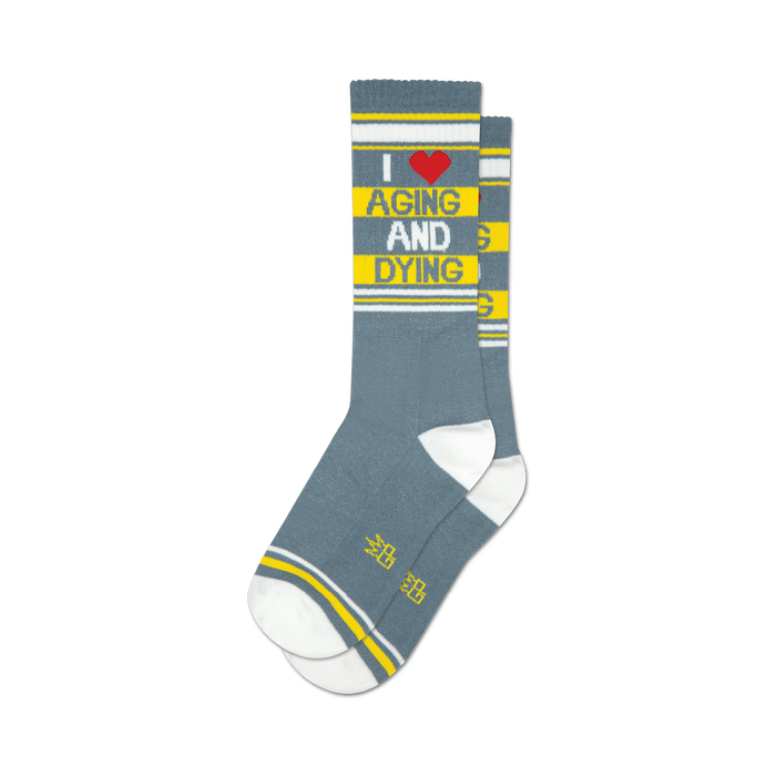 a pair of gray socks with white toes, heels, and cuffs. the socks have red and yellow stripes near the top and yellow stripes above the heel. the words 'i heart aging and dying' are written vertically on the front of the socks in red and yellow letters. }}