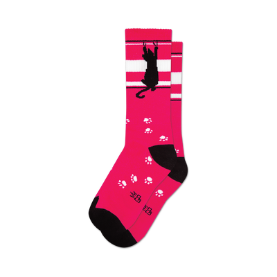 socks that are pink with a black cat and white stripes near the top. the black cat is hanging from the white stripes with one paw and has a paw print by its other paw. there are also paw prints all over the socks.