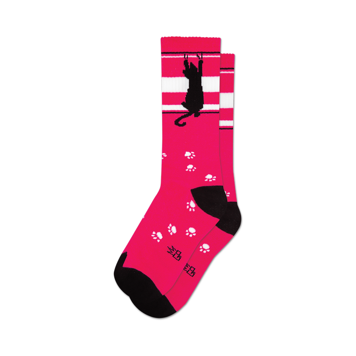 socks that are pink with a black cat and white stripes near the top. the black cat is hanging from the white stripes with one paw and has a paw print by its other paw. there are also paw prints all over the socks. }}