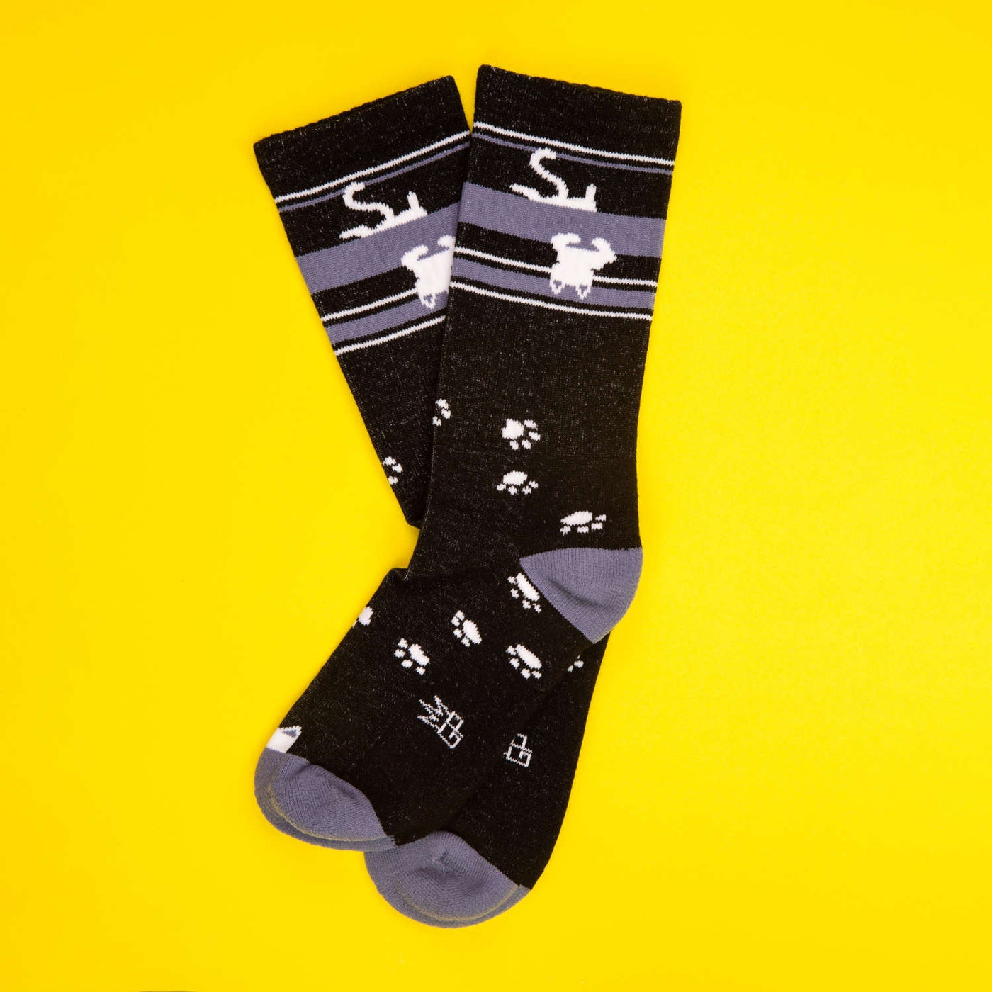 Flatlay product shot of a pair of black crew socks with upside down white cats and paw prints on a yellow background. [lf]