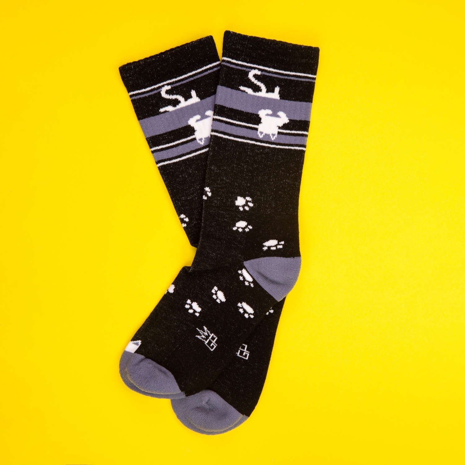 Flatlay product shot of a pair of black crew socks with upside down white cats and paw prints on a yellow background. [lf]