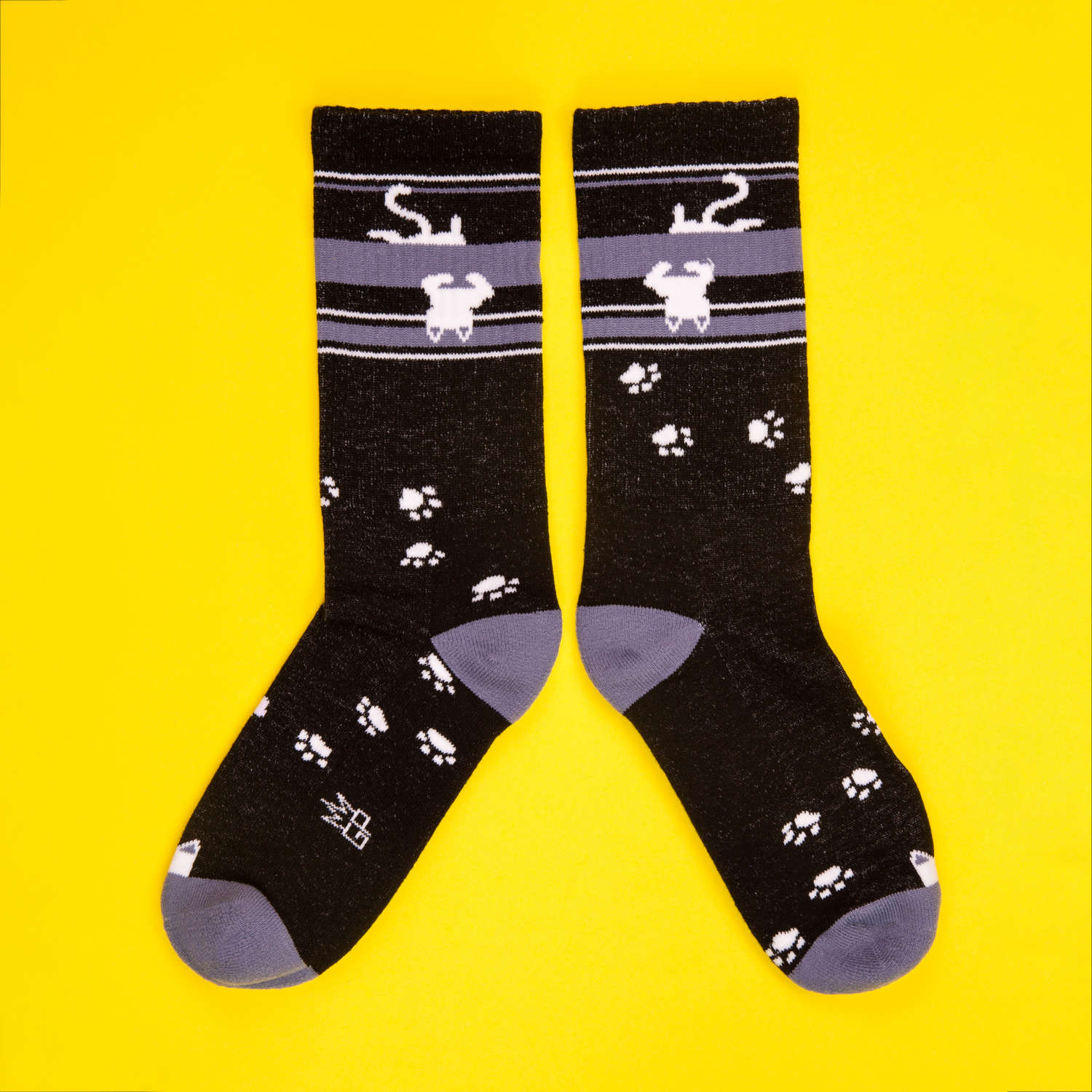 Pair of black crew socks with upside-down white cats and paw prints, laid flat on a yellow background. [lf]
