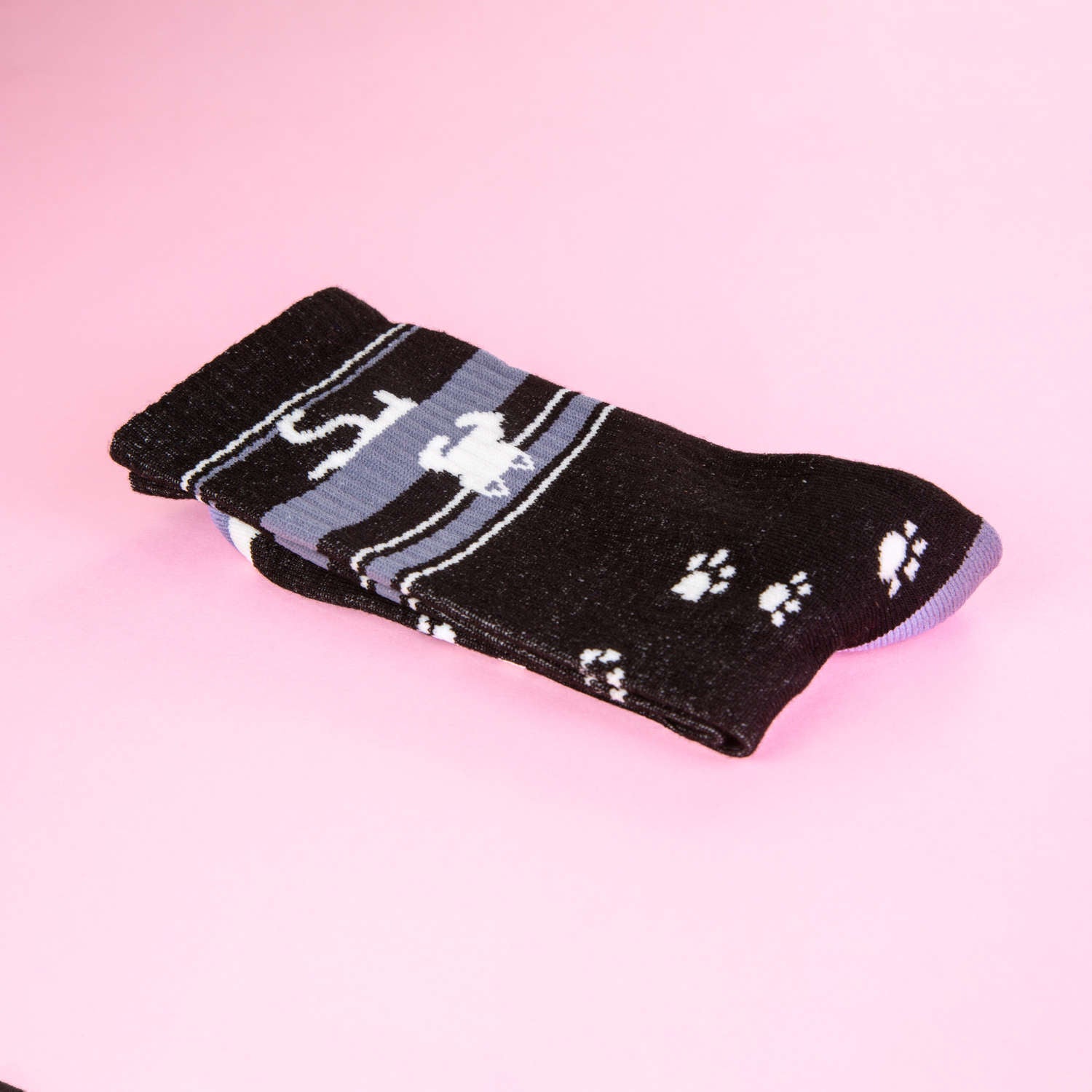 Close-up of a single folded black crew sock with an upside-down white cat and paw print design, on a pink background. [lf]