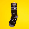 Black crew sock with an upside-down white cat and paw prints hanging on a yellow background with tag. [lf]