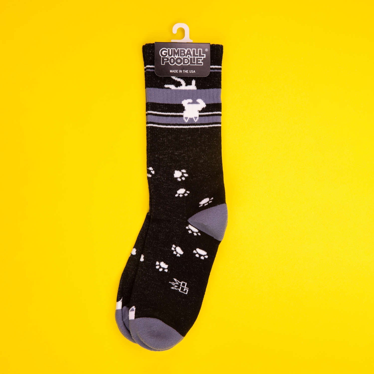 Black crew sock with an upside-down white cat and paw prints hanging on a yellow background with tag. [lf]