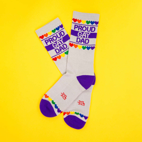 Pair of 'Proud Gay Dad' socks on a yellow background, displayed in an X shape. [lf]