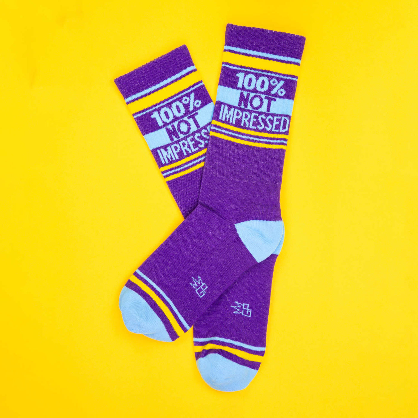 Flat lay of two purple '100% Not Impressed' socks on a yellow background. [lf]