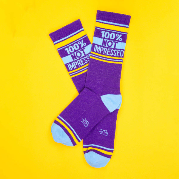 Flat lay of two purple '100% Not Impressed' socks on a yellow background. [lf]