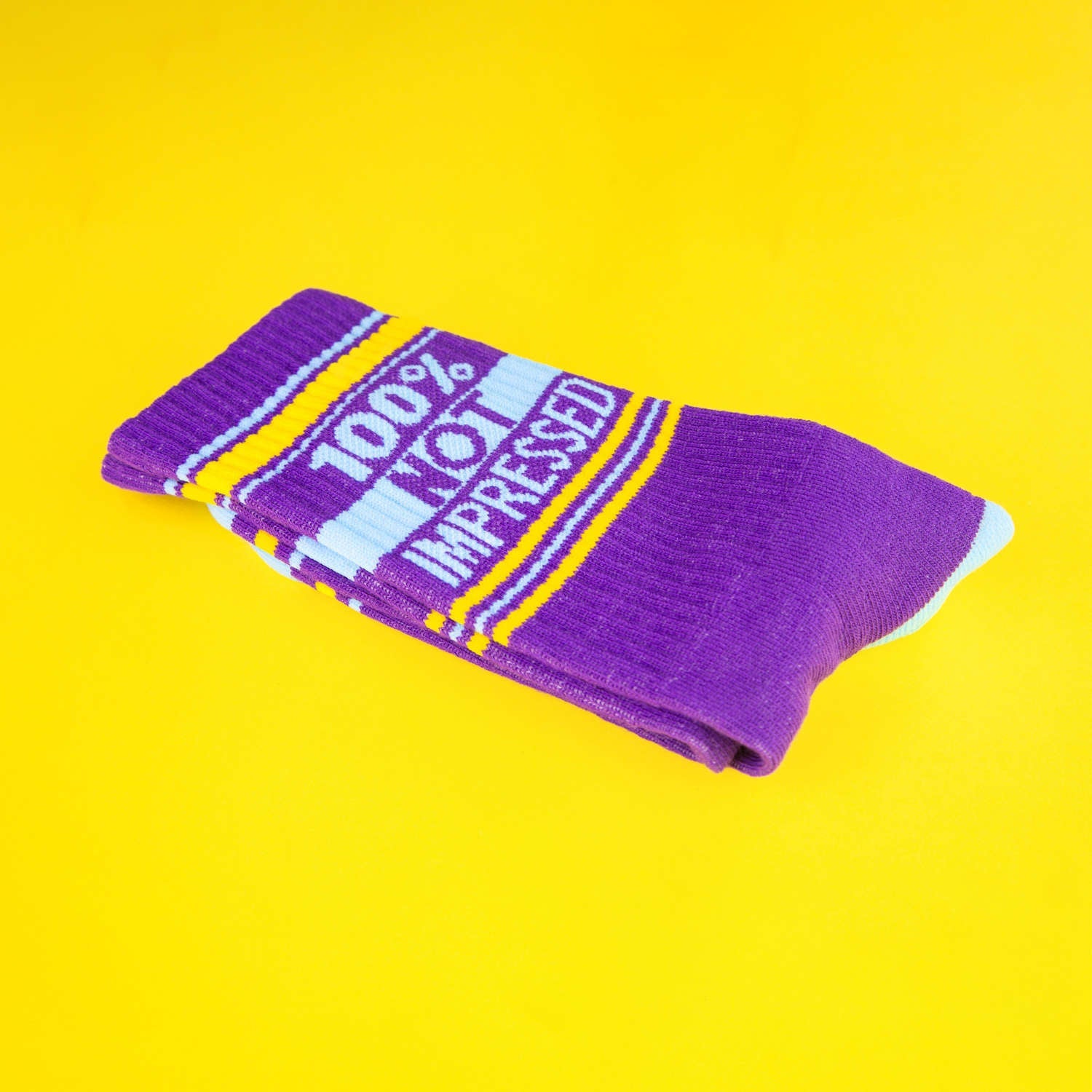 Close-up of a single purple '100% Not Impressed' sock folded on a yellow background. [lf]