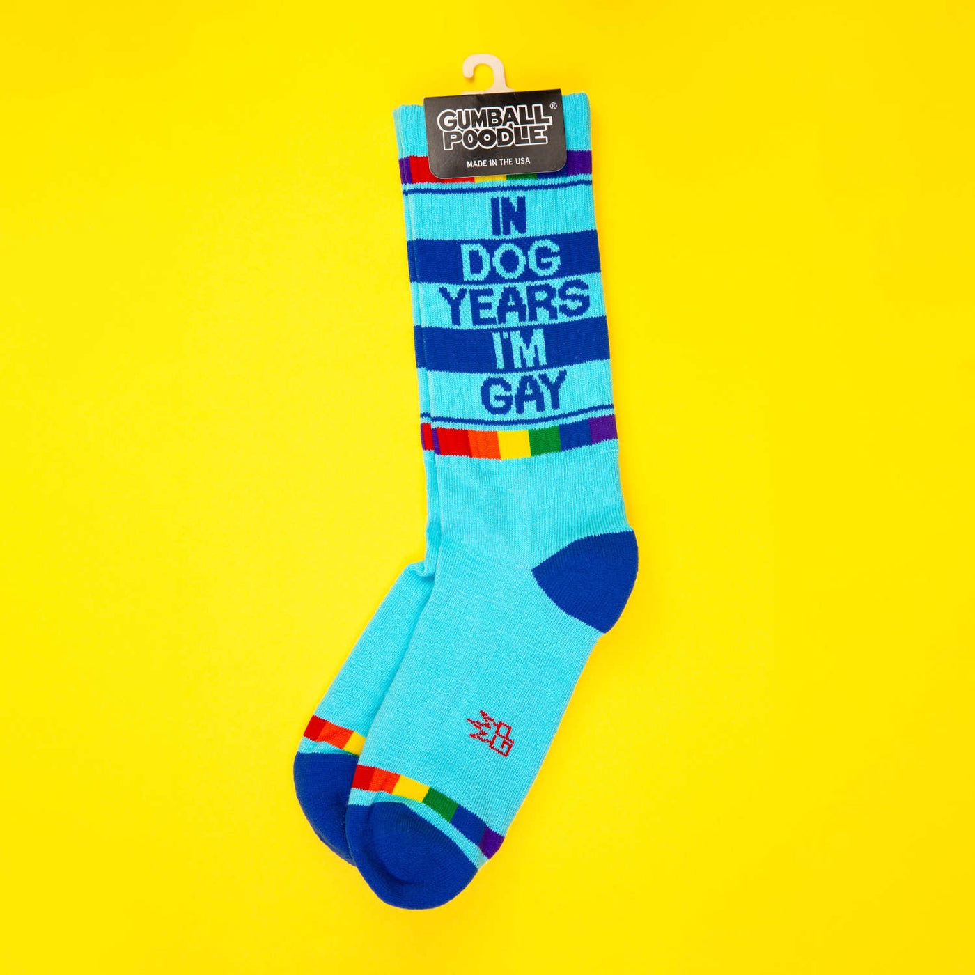Product shot of 'In Dog Years I'm Gay' socks on a yellow background; socks are still on the packaging. [lf]
